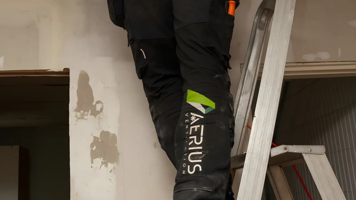 Ventilationsmontör står på en stege framför en öppning i taket i Stockholm med Aerius logotyp på arbetsbyxan