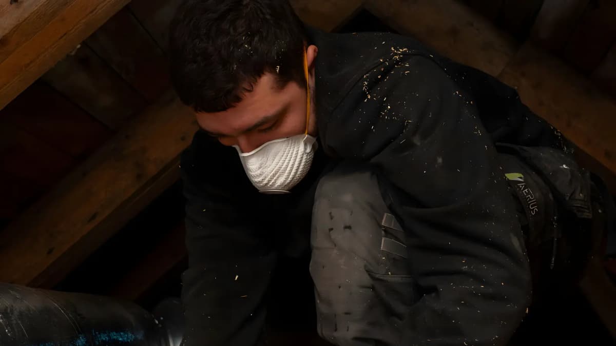 Tekniker med andningsskydd böjer sig ner mot en isolerad ventilationskanal på en mörk vind i Stockholm
